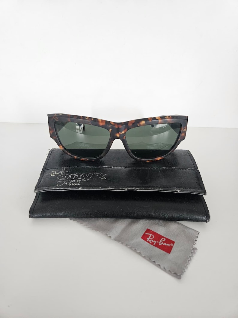 May include: Tortoiseshell-patterned sunglasses with green lenses rest on a black leather case. A gray cleaning cloth with a red Ray-Ban logo is below. The sunglasses have a distinctive, angular frame design.