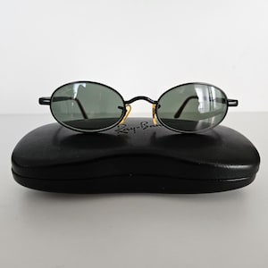 Puede incluir: Gafas de sol ovaladas negras con lentes verdes sobre un estuche negro. Las gafas tienen una montura fina negra con detalles dorados en las bisagras. El estuche tiene forma rectangular redondeada.