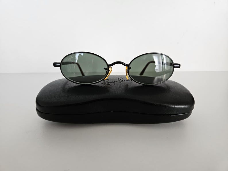 Puede incluir: Gafas de sol ovaladas negras con lentes verdes sobre un estuche negro. Las gafas tienen una montura fina negra con detalles dorados en las bisagras. El estuche tiene forma rectangular redondeada.