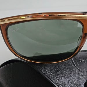 Pu&ograve; includere: Occhiali da sole marroni con barra frontale e dettagli della montatura in metallo dorato. Le lenti sono verde scuro e presentano il logo Ray-Ban stampato in bianco nell'angolo in alto a sinistra. Gli occhiali poggiano su una custodia in pelle nera.