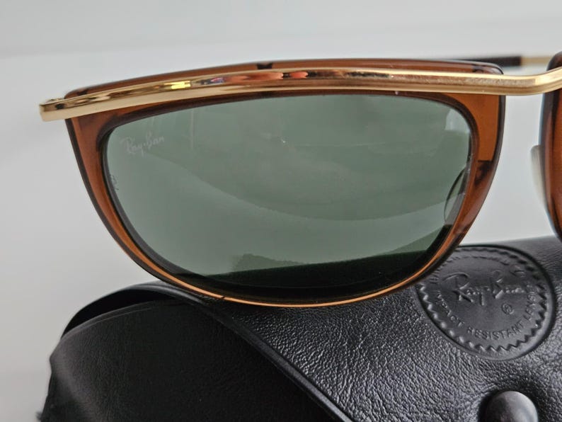 Pu&ograve; includere: Occhiali da sole marroni con barra frontale e dettagli della montatura in metallo dorato. Le lenti sono verde scuro e presentano il logo Ray-Ban stampato in bianco nell'angolo in alto a sinistra. Gli occhiali poggiano su una custodia in pelle nera.