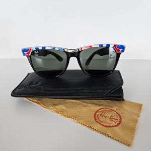 May include: Black Ray-Ban sunglasses with a colourful pattern on the top of the frames. The sunglasses rest on a black case and a tan cleaning cloth with a red Ray-Ban logo.
