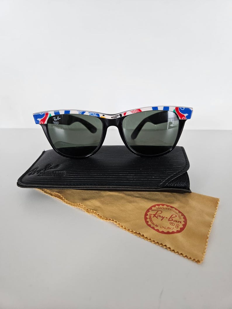 May include: Black Ray-Ban sunglasses with a colourful pattern on the top of the frames. The sunglasses rest on a black case and a tan cleaning cloth with a red Ray-Ban logo.