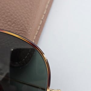 Puede incluir: Primer plano de gafas de sol con montura carey y lentes verdes. Las bisagras y los detalles dorados de la montura a&ntilde;aden un toque de elegancia. Un estuche de cuero marr&oacute;n claro es parcialmente visible en el fondo.