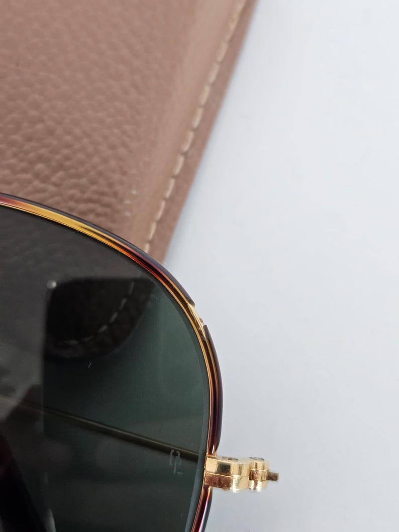 Puede incluir: Primer plano de gafas de sol con montura carey y lentes verdes. Las bisagras y los detalles dorados de la montura a&ntilde;aden un toque de elegancia. Un estuche de cuero marr&oacute;n claro es parcialmente visible en el fondo.
