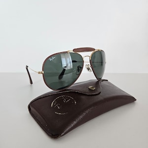 Puede incluir: Gafas de sol estilo aviador con puente de cuero marr&oacute;n y montura de metal dorado. Las lentes son de color verde oscuro y las gafas descansan sobre un estuche de cuero marr&oacute;n. El logotipo de Ray-Ban es visible.