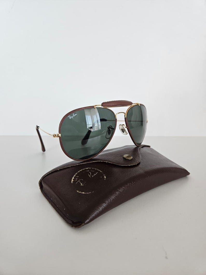 Puede incluir: Gafas de sol estilo aviador con puente de cuero marr&oacute;n y montura de metal dorado. Las lentes son de color verde oscuro y las gafas descansan sobre un estuche de cuero marr&oacute;n. El logotipo de Ray-Ban es visible.