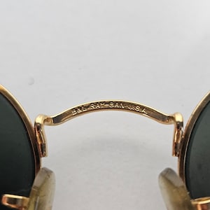 May include: Close-up of gold-coloured sunglasses with dark green lenses. The bridge of the glasses is inscribed with "B&L RAY-BAN U.S.A." The sunglasses have a classic, round shape.