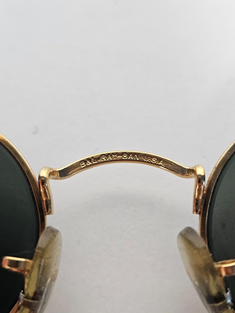 May include: Close-up of gold-coloured sunglasses with dark green lenses. The bridge of the glasses is inscribed with "B&L RAY-BAN U.S.A." The sunglasses have a classic, round shape.