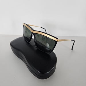 Puede incluir: Gafas de sol negras con detalles dorados y lentes oscuras sobre un estuche negro. Las gafas tienen una montura rectangular con un borde dorado en la parte superior y las patillas. El estuche es de forma ovalada.