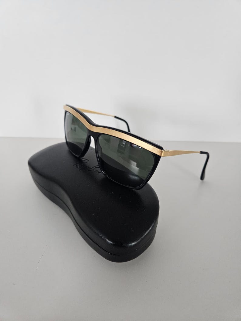 Puede incluir: Gafas de sol negras con detalles dorados y lentes oscuras sobre un estuche negro. Las gafas tienen una montura rectangular con un borde dorado en la parte superior y las patillas. El estuche es de forma ovalada.