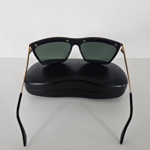 Puede incluir: Gafas de sol negras con patillas doradas y lentes verde oscuro sobre un estuche negro. Las gafas de sol tienen forma rectangular con bordes redondeados y peque&ntilde;os remaches plateados en la montura.