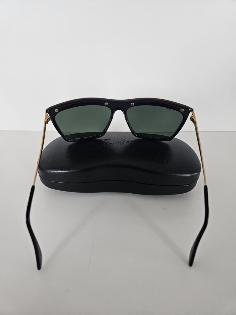 Puede incluir: Gafas de sol negras con patillas doradas y lentes verde oscuro sobre un estuche negro. Las gafas de sol tienen forma rectangular con bordes redondeados y peque&ntilde;os remaches plateados en la montura.