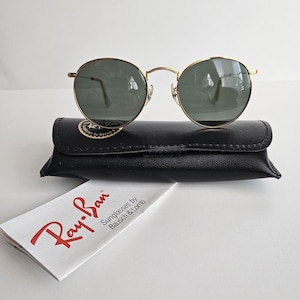 May include: Gold-framed sunglasses with green lenses rest on a black leather case. A white paper with the red "Ray-Ban" logo and text "Sunglasses by Bausch & Lomb" is in front of the case. The sunglasses have a classic, round shape.