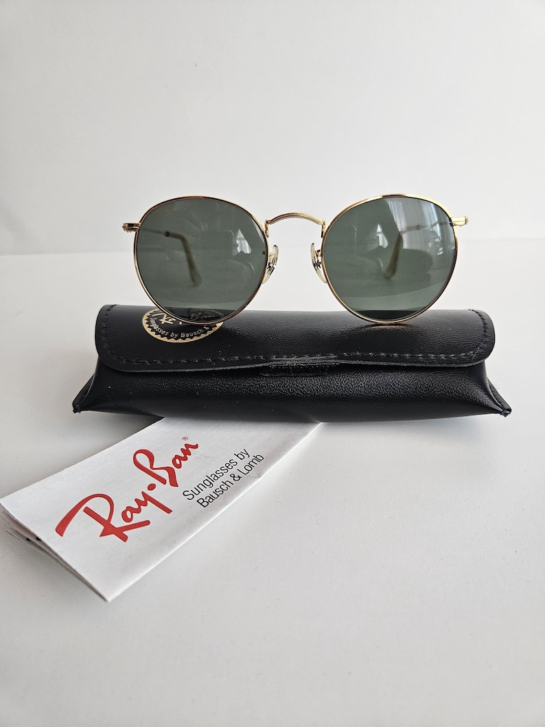 May include: Gold-framed sunglasses with green lenses rest on a black leather case. A white paper with the red "Ray-Ban" logo and text "Sunglasses by Bausch & Lomb" is in front of the case. The sunglasses have a classic, round shape.
