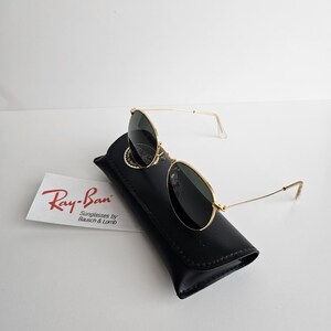 May include: Gold-framed Ray-Ban sunglasses with dark lenses rest on a black leather case. A white card with the Ray-Ban logo and text "Sunglasses by Bausch & Lomb" is visible. The sunglasses have a classic aviator design.
