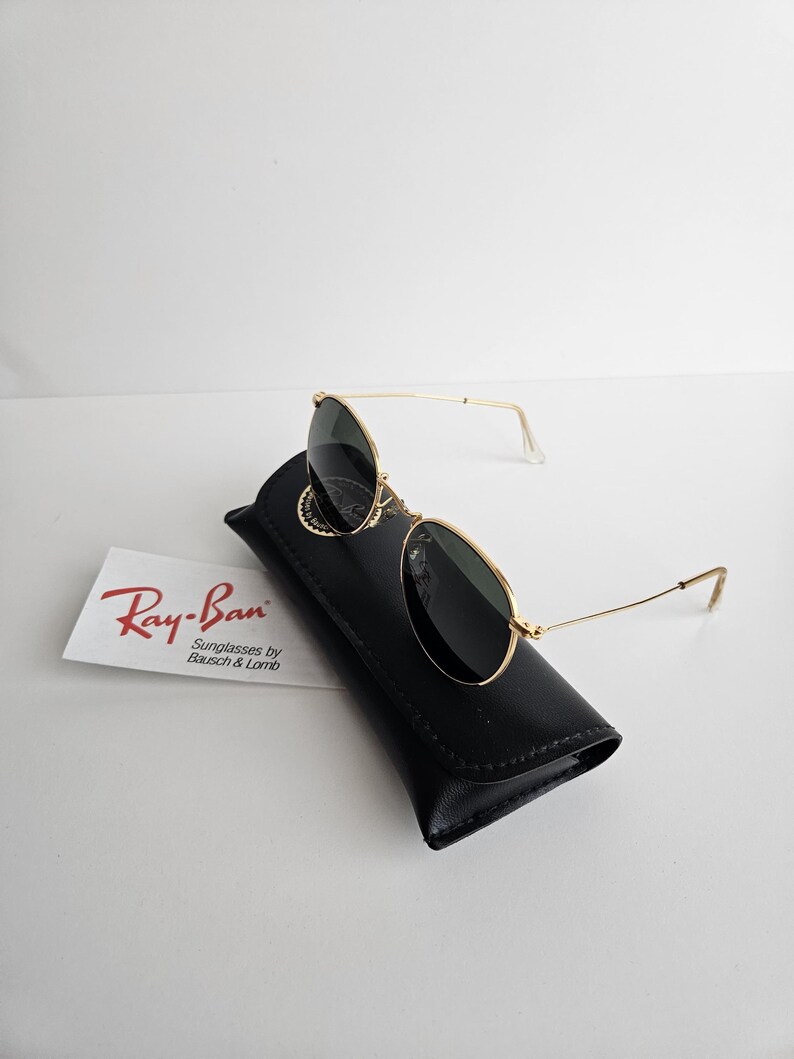 May include: Gold-framed Ray-Ban sunglasses with dark lenses rest on a black leather case. A white card with the Ray-Ban logo and text "Sunglasses by Bausch & Lomb" is visible. The sunglasses have a classic aviator design.