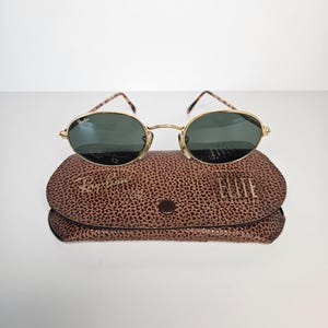 May include: Gold-rimmed sunglasses with oval lenses sit on a brown textured case. The sunglasses have dark green lenses and patterned arms. The case is closed with a small button and features the brand name.