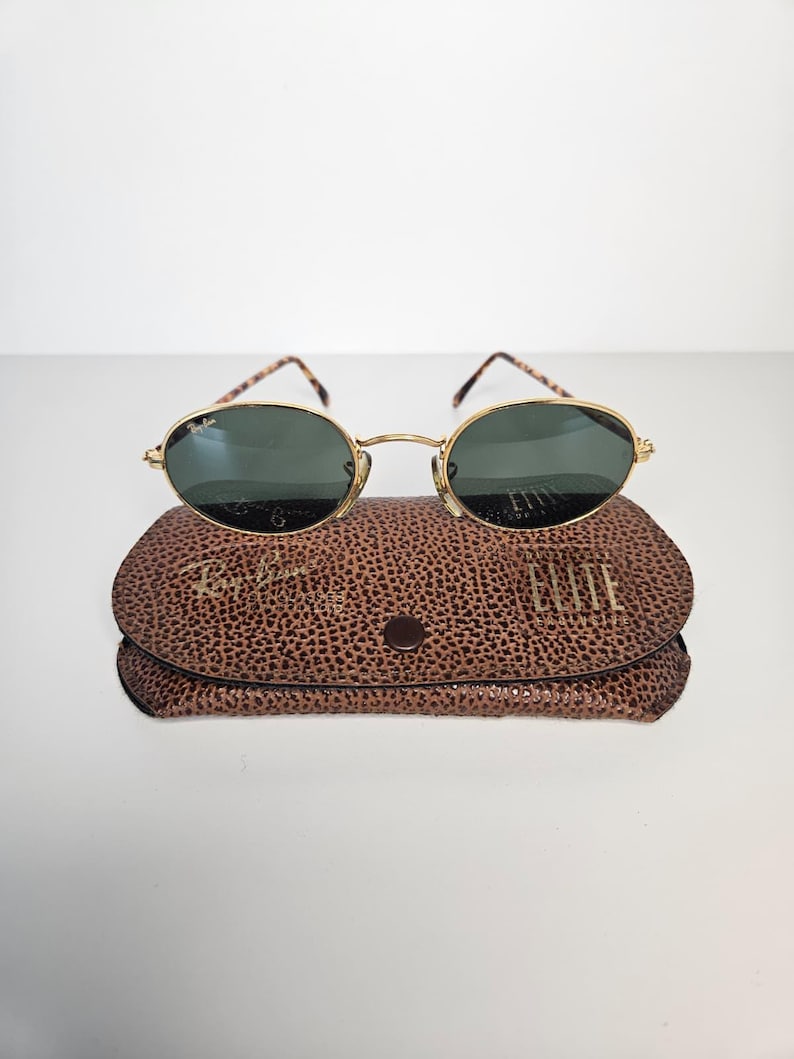 May include: Gold-rimmed sunglasses with oval lenses sit on a brown textured case. The sunglasses have dark green lenses and patterned arms. The case is closed with a small button and features the brand name.