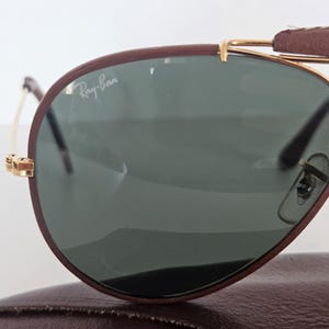 Puede incluir: Primer plano de gafas de sol Ray-Ban con montura de cuero marr&oacute;n y detalles met&aacute;licos dorados. Las lentes son de color verde oscuro. El logotipo de Ray-Ban es visible en la lente. Las gafas de sol descansan sobre un estuche de cuero marr&oacute;n.