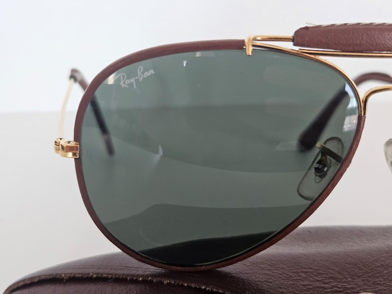 Puede incluir: Primer plano de gafas de sol Ray-Ban con montura de cuero marr&oacute;n y detalles met&aacute;licos dorados. Las lentes son de color verde oscuro. El logotipo de Ray-Ban es visible en la lente. Las gafas de sol descansan sobre un estuche de cuero marr&oacute;n.