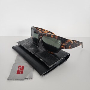 May include: Sunglasses with a tortoiseshell pattern frame and dark lenses rest on a black leather case. A gray cleaning cloth with the Ray-Ban logo is next to the case. The sunglasses are a classic accessory.