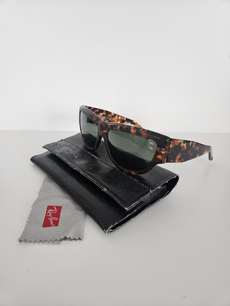 May include: Sunglasses with a tortoiseshell pattern frame and dark lenses rest on a black leather case. A gray cleaning cloth with the Ray-Ban logo is next to the case. The sunglasses are a classic accessory.