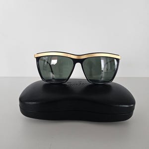 Puede incluir: Gafas de sol negras con montura superior dorada y lentes oscuras, sobre un estuche negro. Las gafas tienen forma rectangular y un dise&ntilde;o cl&aacute;sico. El estuche ovalado tiene el nombre de la marca.