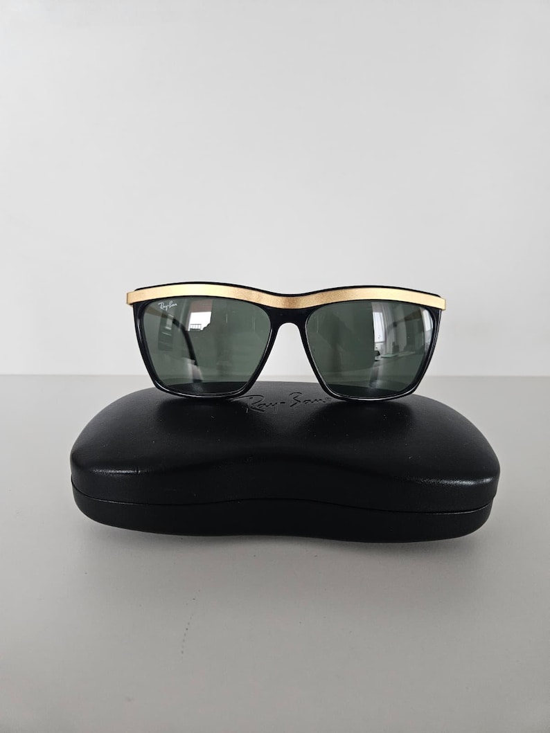 Puede incluir: Gafas de sol negras con montura superior dorada y lentes oscuras, sobre un estuche negro. Las gafas tienen forma rectangular y un dise&ntilde;o cl&aacute;sico. El estuche ovalado tiene el nombre de la marca.
