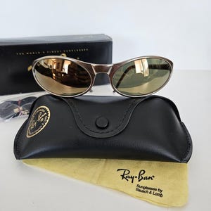 May include: Silver-framed sunglasses with mirrored lenses rest on a black leather case and a yellow cleaning cloth. The case has a button closure and a gold logo. The cloth displays the Ray-Ban logo.