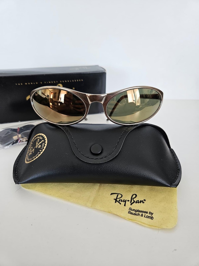 May include: Silver-framed sunglasses with mirrored lenses rest on a black leather case and a yellow cleaning cloth. The case has a button closure and a gold logo. The cloth displays the Ray-Ban logo.