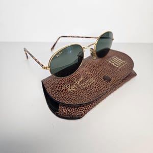 May include: Oval-shaped sunglasses with gold frames and dark lenses, featuring a tortoiseshell pattern on the arms. They sit on a brown leather case, embossed with the Ray-Ban logo and the word "Elite".