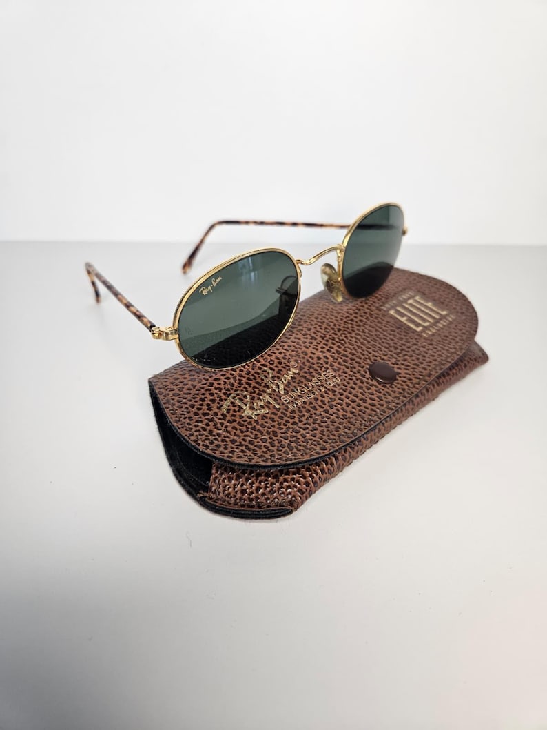 May include: Oval-shaped sunglasses with gold frames and dark lenses, featuring a tortoiseshell pattern on the arms. They sit on a brown leather case, embossed with the Ray-Ban logo and the word "Elite".