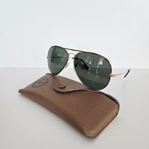 Puede incluir: Gafas de sol aviador con montura dorada y lentes verde oscuro sobre un estuche de cuero marr&oacute;n. El estuche tiene un cierre a presi&oacute;n y un logotipo en relieve. Las gafas de sol son de estilo cl&aacute;sico.