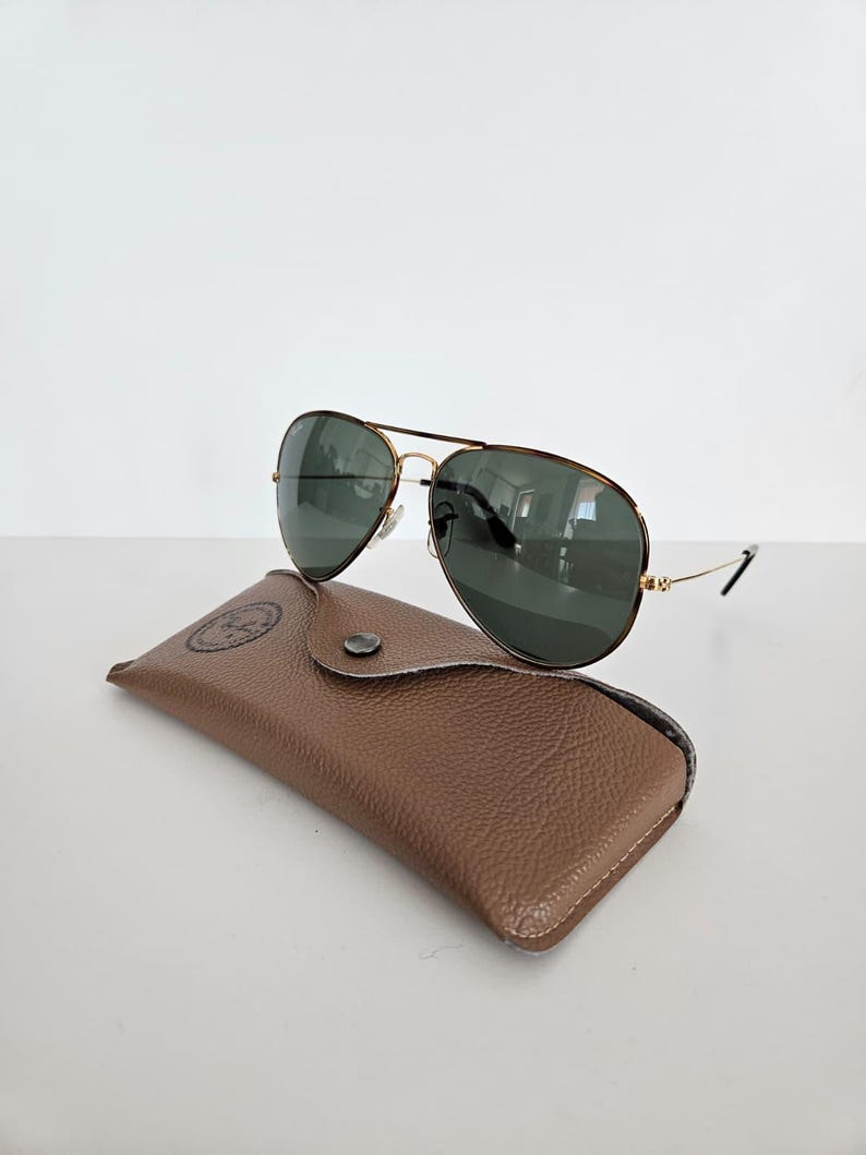 Puede incluir: Gafas de sol aviador con montura dorada y lentes verde oscuro sobre un estuche de cuero marr&oacute;n. El estuche tiene un cierre a presi&oacute;n y un logotipo en relieve. Las gafas de sol son de estilo cl&aacute;sico.
