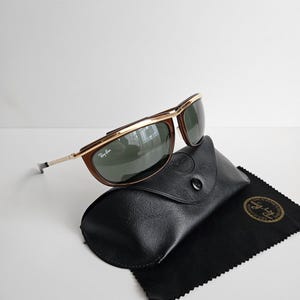 Pu&ograve; includere: Occhiali da sole Ray-Ban con barra frontale e dettagli della montatura in metallo dorato, bordi marroni e lenti verde scuro. Gli occhiali sono posizionati su una custodia nera dall'aspetto in pelle testurizzata con un panno di pulizia nero.
