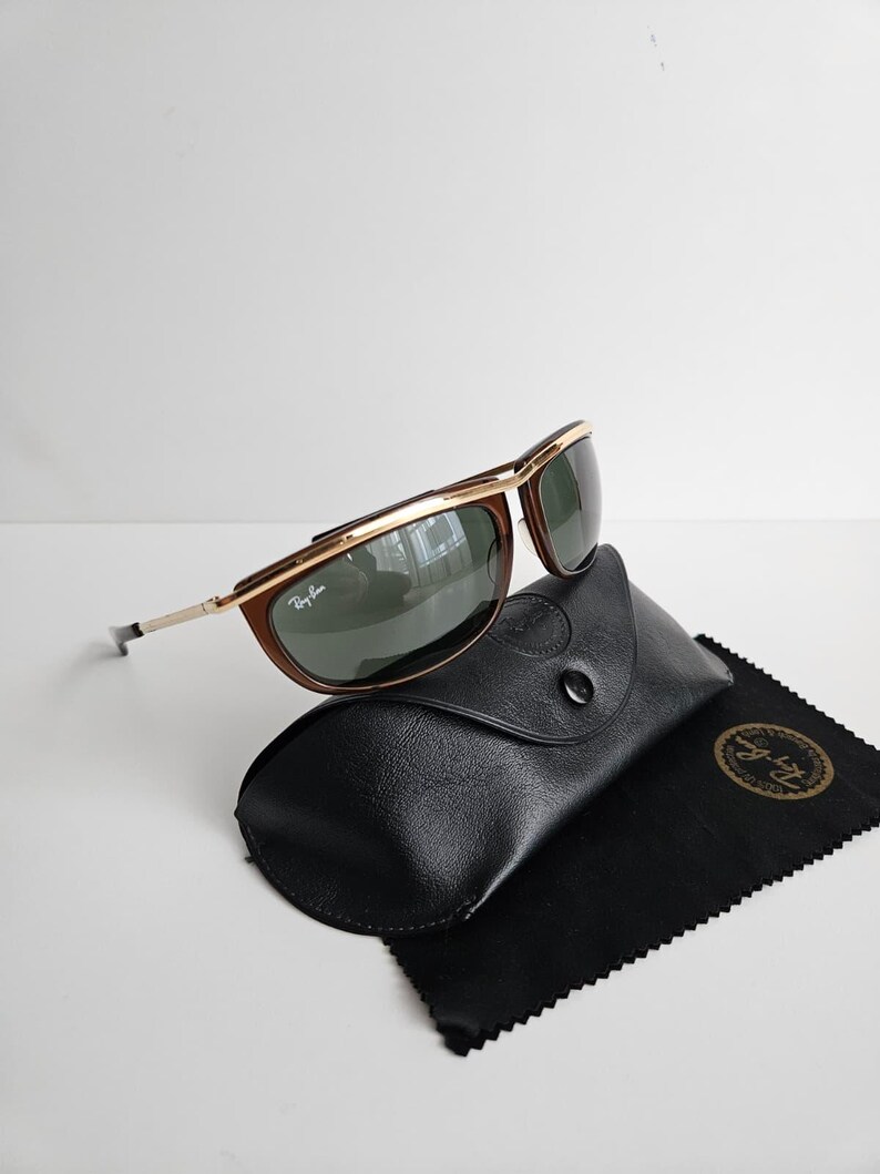 Pu&ograve; includere: Occhiali da sole Ray-Ban con barra frontale e dettagli della montatura in metallo dorato, bordi marroni e lenti verde scuro. Gli occhiali sono posizionati su una custodia nera dall'aspetto in pelle testurizzata con un panno di pulizia nero.