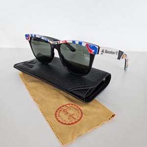 May include: Black sunglasses with a patterned frame, on a black case with a tan cleaning cloth. The frames have the text "Barcelona 92". The cleaning cloth has the "Ray-Ban" logo.