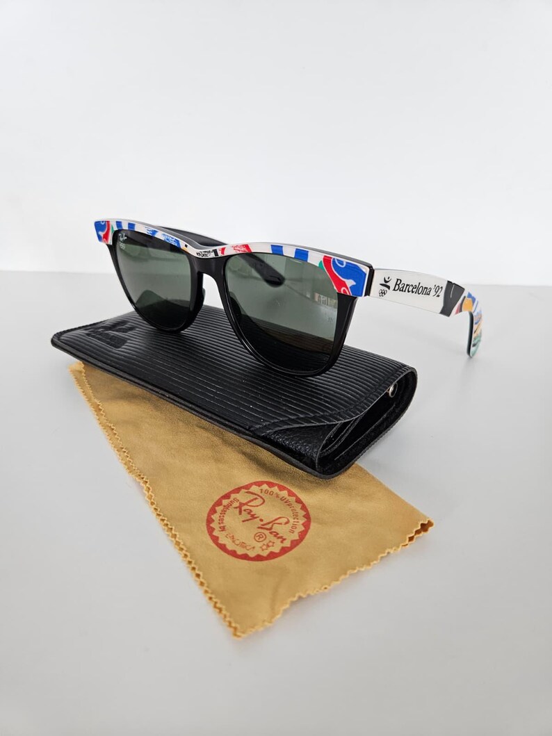 May include: Black sunglasses with a patterned frame, on a black case with a tan cleaning cloth. The frames have the text "Barcelona 92". The cleaning cloth has the "Ray-Ban" logo.