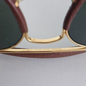 Puede incluir: Primer plano de gafas de sol Ray-Ban con montura dorada y lentes verde oscuro. La barra superior y las patillas son marrones. El texto "B&L RAY-BAN 62 14" est&aacute; impreso en la montura.