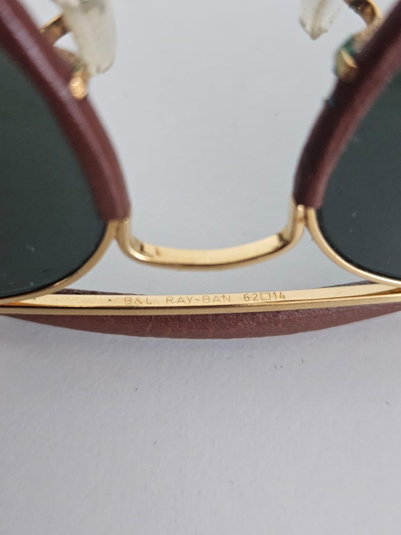 Puede incluir: Primer plano de gafas de sol Ray-Ban con montura dorada y lentes verde oscuro. La barra superior y las patillas son marrones. El texto "B&L RAY-BAN 62 14" est&aacute; impreso en la montura.