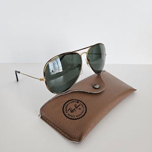 Puede incluir: Gafas de sol aviador con montura dorada y lentes verde oscuro, sobre un estuche de cuero marr&oacute;n. El estuche tiene un logotipo circular. Las gafas de sol tienen un dise&ntilde;o cl&aacute;sico, adecuado para varios estilos.