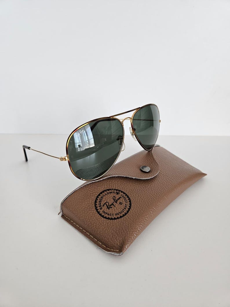 Puede incluir: Gafas de sol aviador con montura dorada y lentes verde oscuro, sobre un estuche de cuero marr&oacute;n. El estuche tiene un logotipo circular. Las gafas de sol tienen un dise&ntilde;o cl&aacute;sico, adecuado para varios estilos.