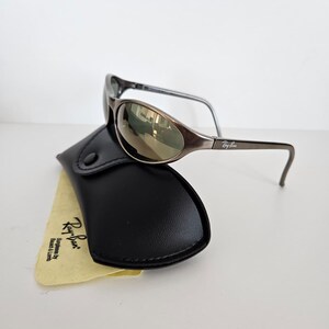 May include: Brown oval sunglasses with a black case and a yellow cleaning cloth. The sunglasses have a metallic frame and mirrored lenses. The case is made of black leather. The cleaning cloth has the Ray-Ban logo.