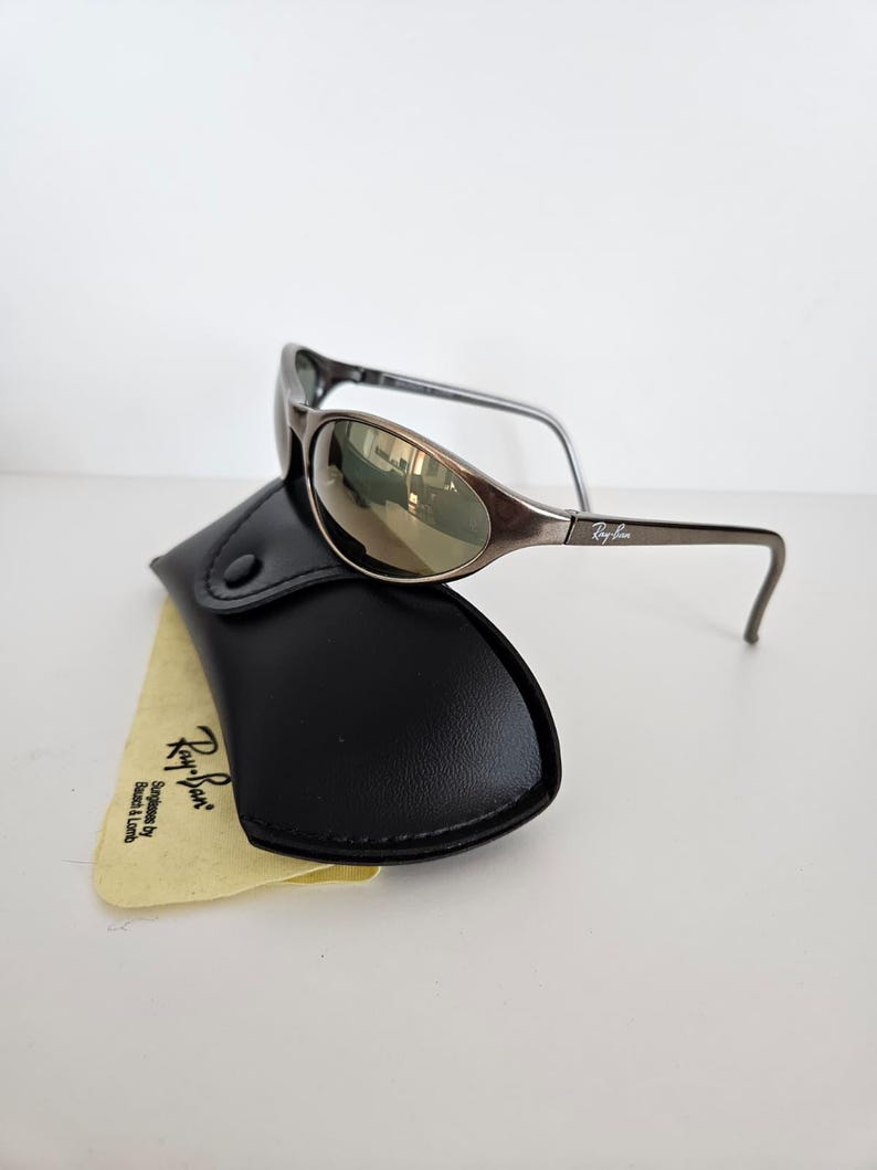 May include: Brown oval sunglasses with a black case and a yellow cleaning cloth. The sunglasses have a metallic frame and mirrored lenses. The case is made of black leather. The cleaning cloth has the Ray-Ban logo.