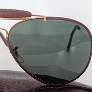 Puede incluir: Gafas de sol estilo aviador con una barra frontal y montura de cuero marr&oacute;n. Las lentes son de color verde oscuro y las monturas son doradas. Las gafas de sol est&aacute;n sobre un estuche de cuero marr&oacute;n.