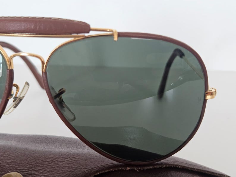 Puede incluir: Gafas de sol estilo aviador con una barra frontal y montura de cuero marr&oacute;n. Las lentes son de color verde oscuro y las monturas son doradas. Las gafas de sol est&aacute;n sobre un estuche de cuero marr&oacute;n.