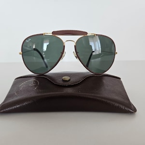 Puede incluir: Gafas de sol estilo aviador con lentes verdes y puente de cuero marr&oacute;n, sobre un estuche de cuero marr&oacute;n. Las gafas tienen una montura dorada y un dise&ntilde;o cl&aacute;sico, ideales para d&iacute;as soleados.