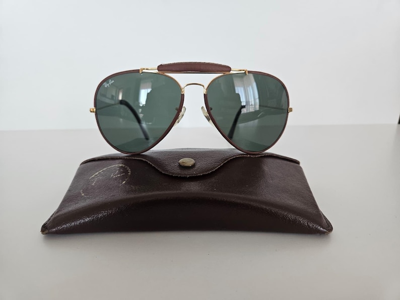 Puede incluir: Gafas de sol estilo aviador con lentes verdes y puente de cuero marr&oacute;n, sobre un estuche de cuero marr&oacute;n. Las gafas tienen una montura dorada y un dise&ntilde;o cl&aacute;sico, ideales para d&iacute;as soleados.