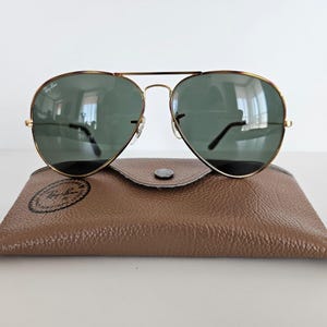 Puede incluir: Gafas de sol aviador con montura dorada y lentes verdes sobre un estuche de cuero marr&oacute;n. El estuche tiene un cierre de bot&oacute;n a presi&oacute;n y un logotipo circular. Las gafas reflejan el entorno.